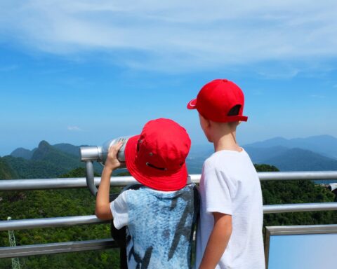 The two boys looks through a telescope. A little boys in a red hats looks through a telescope from a viewing platform on the sea and mountains on a sunny bright day. Langkawi, Malaysia.