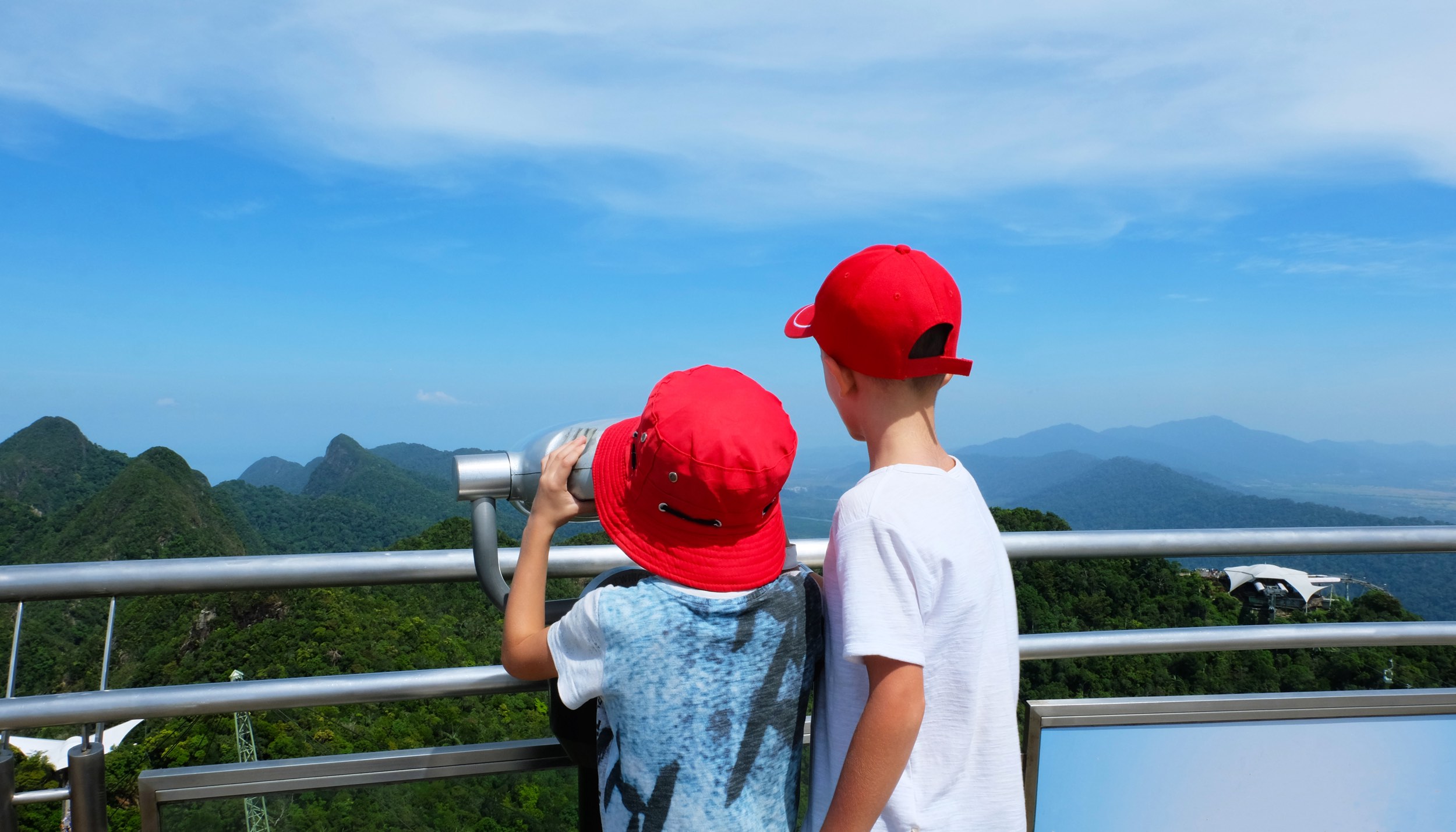 The two boys looks through a telescope. A little boys in a red hats looks through a telescope from a viewing platform on the sea and mountains on a sunny bright day. Langkawi, Malaysia.